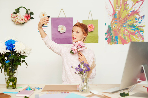 Diseñador floral bastante joven con flores rosadas en la mano tomando selfie contra la pared de estudio colorido decorado con pintura abstracta
 - Foto, Imagen
