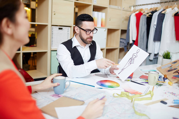 Modern fashion designer pointing at sketch of new dress while consulting with assistant - Photo, Image