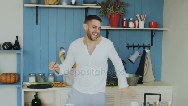Slow motion of Attractive young funny man dancing and singing with ladle while cooking in the kitchen at home - Séquence, vidéo