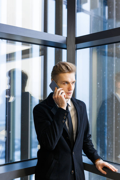 Skoncentrowane młody przedsiębiorca w szykowny garnitur mówi do swojego klienta na smartphone, stojąc przez panoramiczne okna, portret strzał - Zdjęcie, obraz