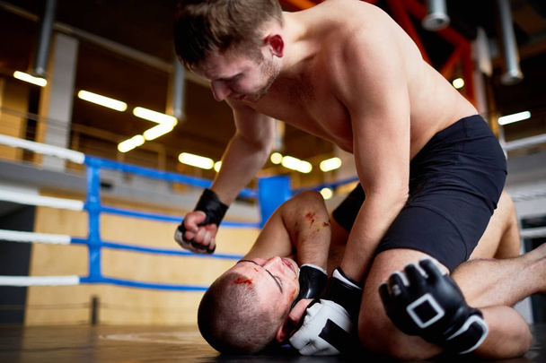 Портрет борцов-профессионалов, сражающихся на боксерском ринге: мужчина бьет кровавого противника в лицо, сталкивая его на пол
 - Фото, изображение