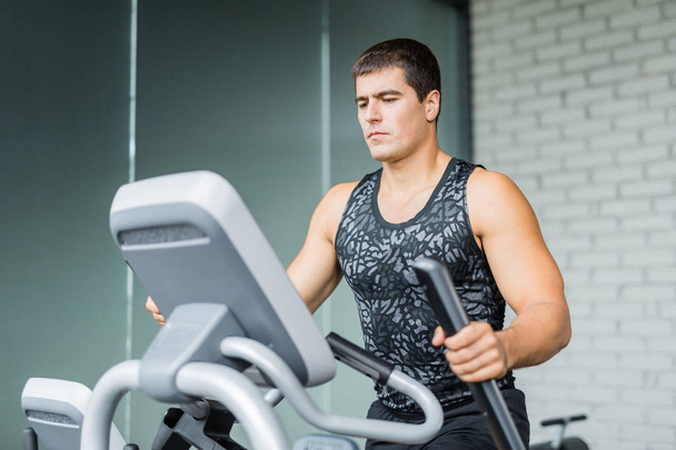 Portrait of muscular sportive man  running using elliptical trainer during workout in modern gym - Photo, Image