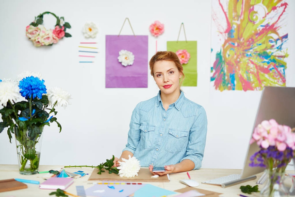 Retrato de cintura hacia arriba del joven diseñador floral confiado mirando a la cámara mientras sostiene el teléfono móvil en la mano, ella sentada en un escritorio desordenado contra la pared decorada con flores y pintura abstracta
 - Foto, imagen