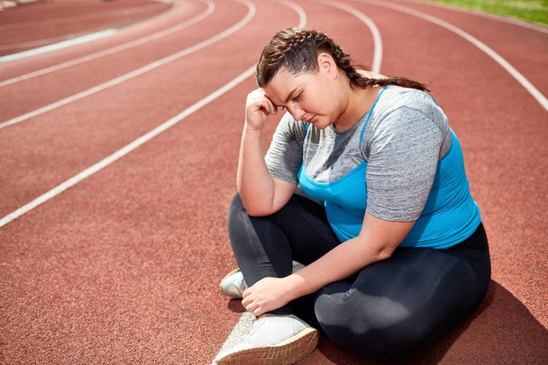 Stressed fat female in activewear leaning on her hand while sitting on racetrack - Foto, imagen