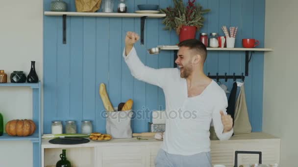 Slow motion of handsome young funny man dancing and singing with ladle while cooking in the kitchen at home - Séquence, vidéo