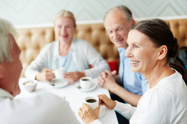 Group of senior companions sitting by table in cafe and having talk by cup of coffee - 写真・画像