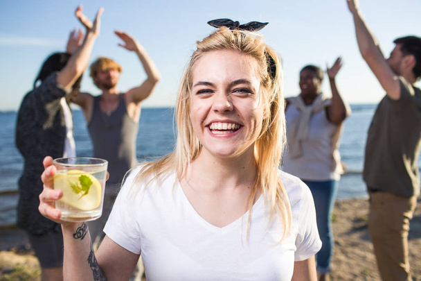 Ecstatic girl with glass of citrus cocktail enjoying gathering with friends on the beach - Photo, Image