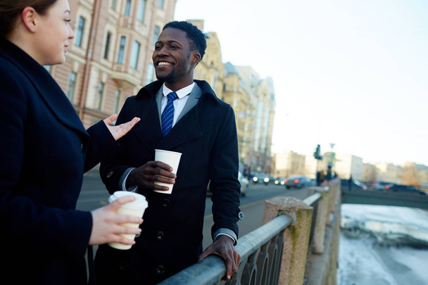 Retrato de dos colegas de negocios, afroamericanos y jóvenes, en la calle de la ciudad por el río hablando entre sí mientras sostienen tazas de café desechables ambos con abrigos
  - Foto, Imagen