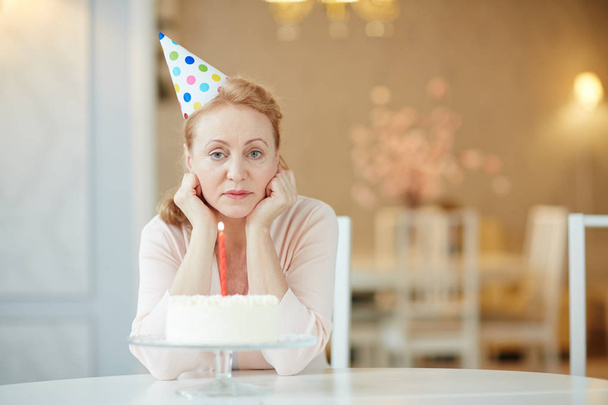 Portrait de triste femme mature solitaire assise seule à la table d'anniversaire avec gâteau, portant un chapeau de fête et regardant la caméra
 - Photo, image
