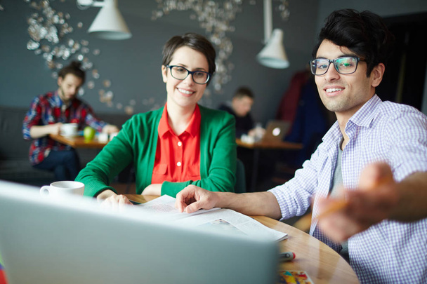 Portret van twee jonge collega's man en vrouw zowel het dragen van casual kleding en bril, bespreken van werk op de bijeenkomst in café en wordt gewezen op de laptop scherm - Foto, afbeelding