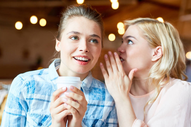 Attractive blond-haired woman sharing secret with friend while sitting at cozy small coffeehouse and enjoying cappuccino, blurred background - Foto, imagen