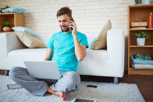 Позитивний молодий чоловік з бородою говорить по телефону, коли він використовує ноутбук у вітальні
 - Фото, зображення