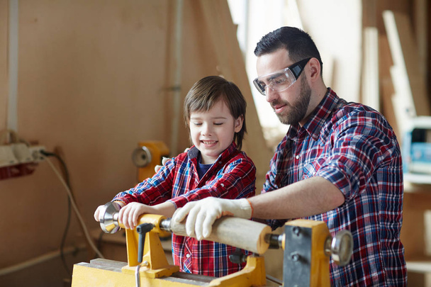 Falegname in occhiali protettivi e suo figlio lavorazione pezzo di legno in officina
 - Foto, immagini