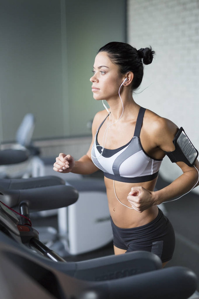 Retrato de mujer morena deportiva haciendo ejercicio en la cinta de correr en el gimnasio escuchando música usando el soporte para smartphone de hombro
 - Foto, imagen