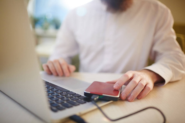 Крупный план неузнаваемого бизнесмена в застегнутой белой рубашке, сидящего за столом кафе и использующего ноутбук, подключенный к внешнему жесткому диску
 - Фото, изображение