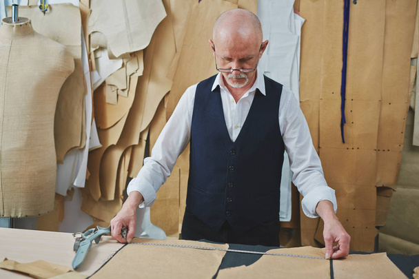 Portrait of skilled senior tailor looking at patterns while making clothes in atelier shop - Photo, Image