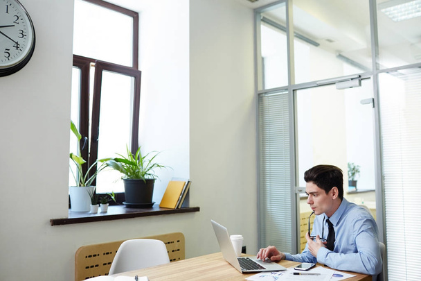 Concentrado jovem trabalhador de colarinho branco sentado na frente do laptop e trabalhando em projeto promissor, interior da sala de reuniões moderna no fundo
 - Foto, Imagem