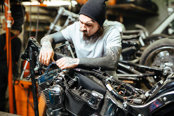 Porträt eines modernen, schwer tätowierten Mannes, der in der Garage bei harter Beleuchtung maßgeschneiderte Motorräder zusammenbaut - Foto, Bild