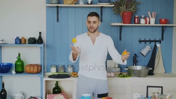Funny cheerful man juggle with orange mandarins it the kitchen at home - Filmati, video