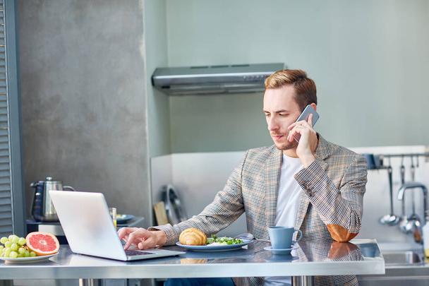 Busy young manager discussing order details with client and using laptop while having breakfast at modern kitchen, waist-up portrait - 写真・画像