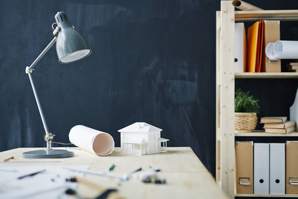 Background shot of architects workplace with house model on desk, metal lamp, blueprints and supplies against blackboard in modern office, copy space - Фото, изображение