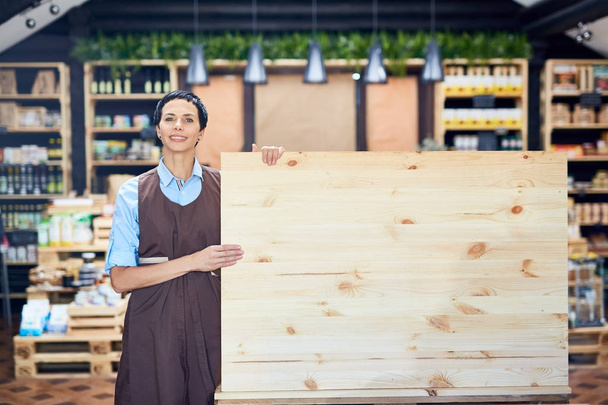 Vrij middelbare leeftijd winkelbediende in schort leeg houten bord terwijl staande te houden in mooie winkel met biologische producten, portret shot - Foto, afbeelding