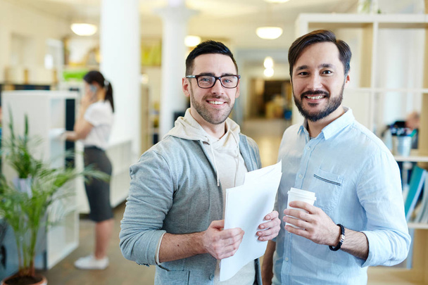 Portrait of two modern professionals looking at camera and smiling standing in open office space - Фото, изображение