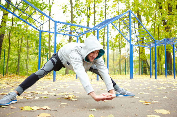 Jovem desportivo fazendo exercícios de alongamento antes de correr, parque de treino de rua com folhas caídas no fundo
 - Foto, Imagem