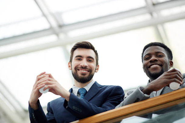 Retrato de bajo ángulo del guapo hombre de negocios barbudo con un colega afroamericano mirando desde el balcón de cristal y sonriendo sosteniendo tazas durante el descanso
 - Foto, imagen