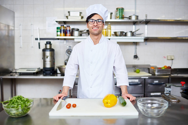Feliz joven chef en uniforme de pie junto al lugar de trabajo en la cocina
 - Foto, Imagen