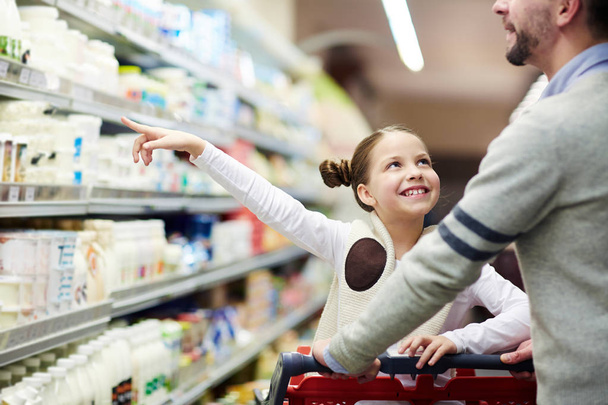 Happy family buying groceries: smiling little girl choosing dairy products from fridge in milk aisle while shopping in supermarket - Фото, изображение