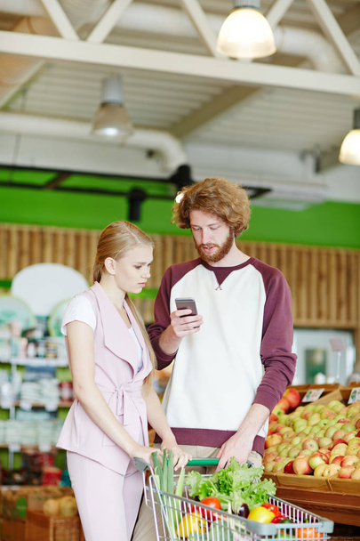 Μοντέρνο ζευγάρι ψάχνει μέσα από το νέο promo σε smartphone σε σούπερ μάρκετ - Φωτογραφία, εικόνα