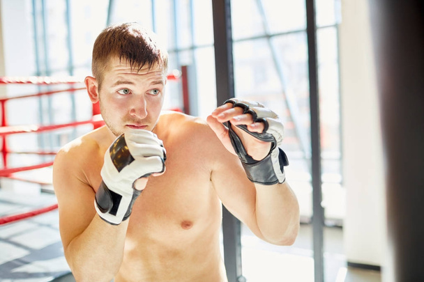 Retrato del sudoroso hombre deportivo sin camisa golpeando saco de boxeo durante la práctica de boxeo en el club de lucha iluminado por el sol
 - Foto, imagen