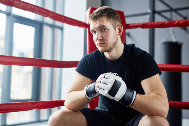 Portrait of young man posing in boxing ring sitting on floor and looking at camera looking at camera with confidence - Foto, afbeelding