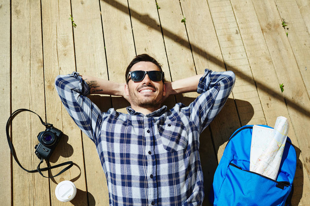 Top vista retrato de belo jovem deitado em pranchas de madeira doca desfrutando de luz solar em viagem turística com câmera fotográfica e mochila nas proximidades
 - Foto, Imagem