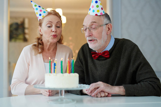 Portrét milující starší pár slaví narozeniny spolu sedí u stolu s dortem a nosí klobouky foukání svíčky a realizací přání - Fotografie, Obrázek