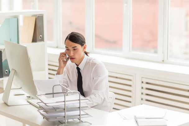 Bilgisayar başında oturan ve sipariş ayrıntıları üstünde hareket eden telefon, zemin üzerine panoramik pencereli ferah ofis iç müşteri ile tartışırken yakışıklı Asya beyaz yakalı işçi - Fotoğraf, Görsel