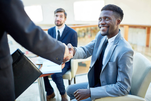 Portrait of smiling young African-American  businessman shaking hands with partner, sitting at table in meeting - Foto, imagen