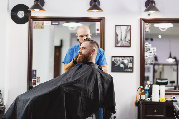 Чоловічий професійний перукар, що обслуговує клієнта по кліпу. Імбир красивий брутальний стильний молодий чоловік з товстою великою бородою і коротким волоссям стає модною зачіскою в чорній накидці. Світло-білий перукарня
 - Фото, зображення