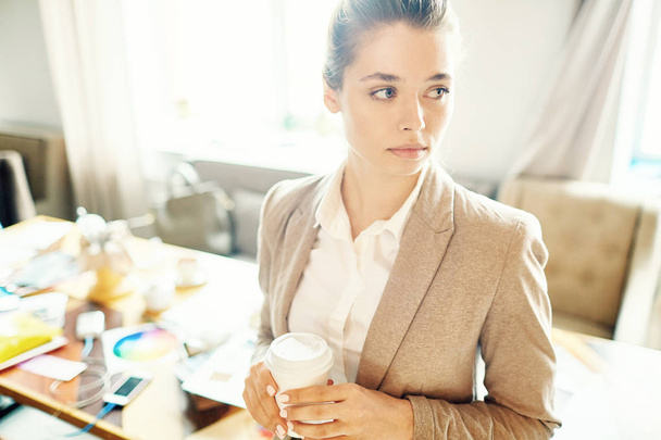 Ελκυστική νεαρή υπαλληλικός εργαζόμενος βλέπει μακριά pensively ενώ στέκεται στο ενιαίο γραφείο με χαρτί Κύπελλο του καφέ στα χέρια, Μέση-κατακόρυφα - Φωτογραφία, εικόνα