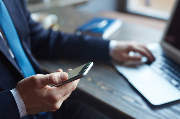 Retrato de close-up de empresário de sucesso: mão masculina segurando smartphone, digitando mensagens e navegando na internet enquanto trabalhava com laptop
 - Foto, Imagem