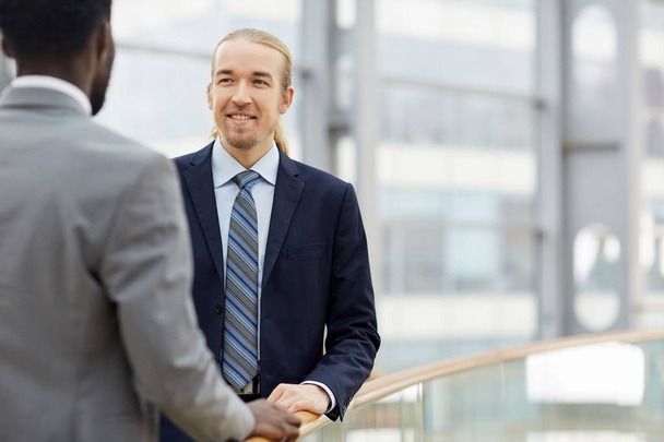 Portrait of two contemporary businessmen, one of them African, talking casually and smiling by glass railing in modern office building - Photo, Image