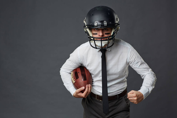 Agresivní podnikatel středního věku s barvou na jeho tvář nosí helmu amerického fotbalu a držení koule při pohledu na fotoaparát proti šedé pozadí - Fotografie, Obrázek
