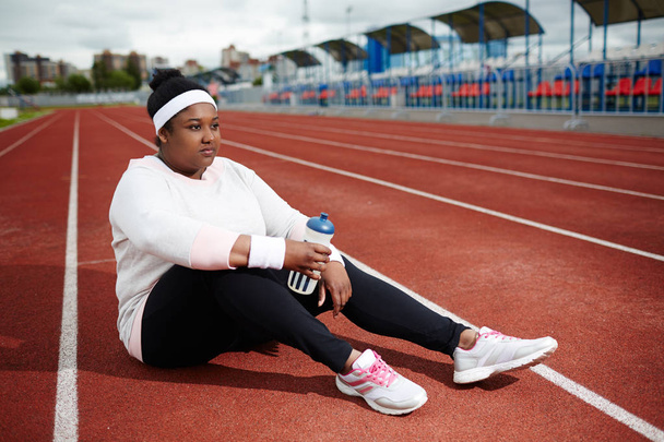 Υπέρβαρα Αφρικανή γυναίκα σε αθλητικών ενδυμάτων συνεδρίαση της τροχιάς, αναρρώνει μετά από τρέξιμο πρακτικής και σκέψης - Φωτογραφία, εικόνα