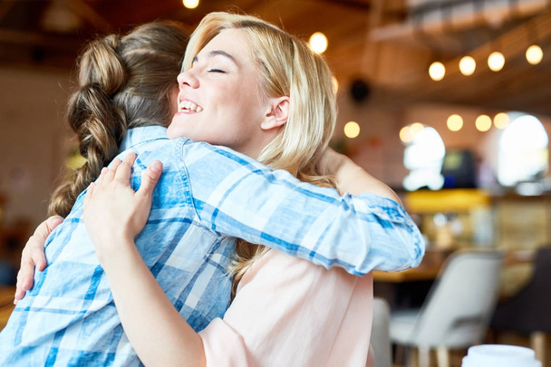 Twee vrolijke en vriendelijke meisjes groeten elkaar door omhelzing in café - Foto, afbeelding