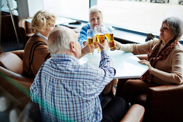 Skupina radostné starších lidí, posezení v prostorné kavárně s panoramatickými okny a opékání s pivní sklenice - Fotografie, Obrázek