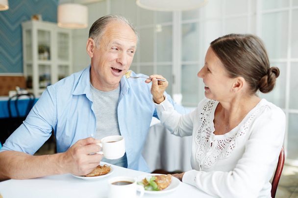 Sorrindo mulher sênior alimentando seu marido bonito com bolo apetitoso ao ter data em adorável pequeno café
 - Foto, Imagem