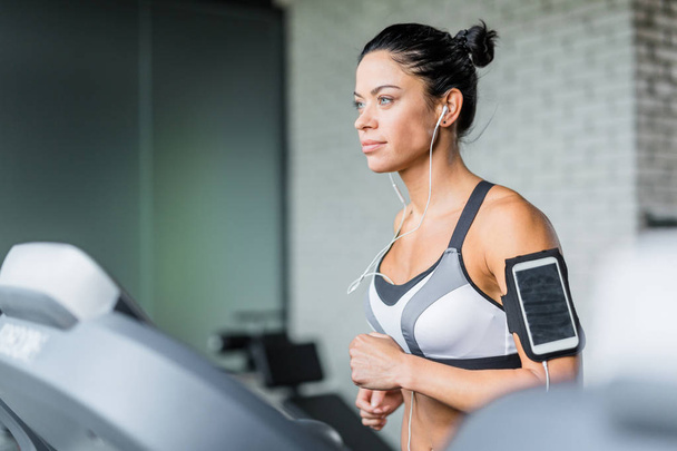 Portrait of sportive brunette woman  running on treadmill listening to music using shoulder smartphone holder during workout in gym - Photo, Image