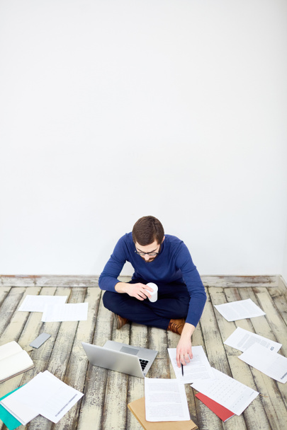 Portrait of  modern freelancer sitting cross legged on floor in office,  working with laptop and  documents laid out around - Фото, зображення