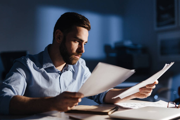 Portrait of focused bearded businessman wearing casual clothes working with documentation in dark room late at night, his face lit by lamp light - Photo, Image